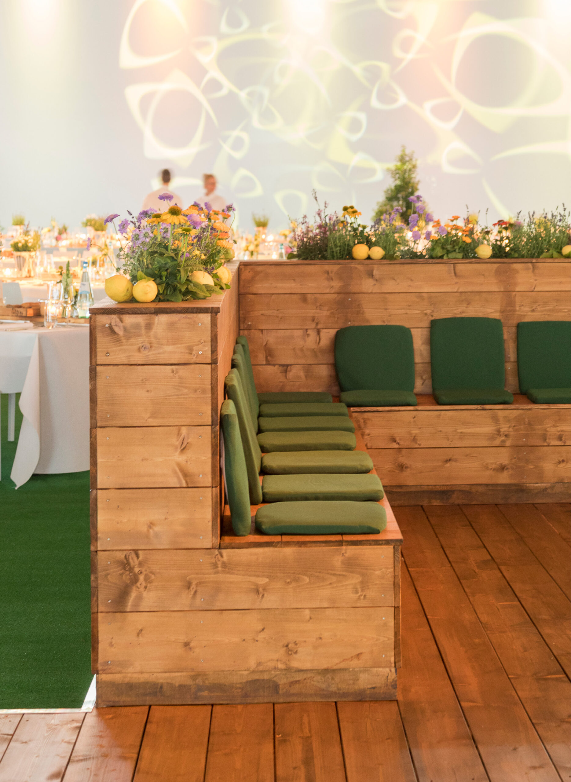 Holzbänke mit grünen Kissen und Blumen, eingebettet in einen hellen, dekorierten Veranstaltungsraum mit Tischen - ideal für Messecatering für Veranstaltungen.