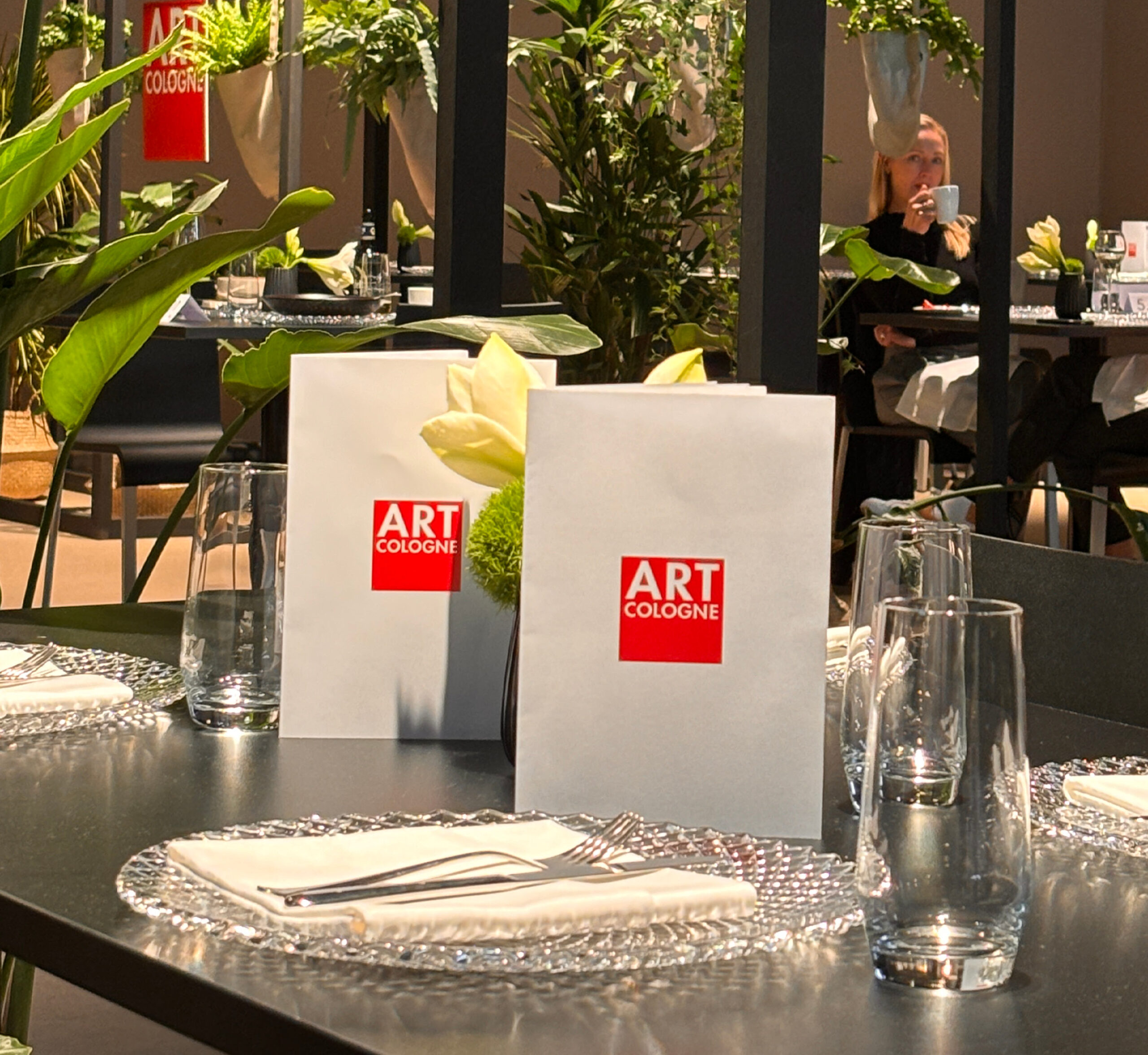 Tischset mit Tellern, Gläsern und Servietten; ART COLOGNE-Menüs und Pflanzen im modernen Restaurantambiente - ideal für Messecatering für Veranstaltungen.