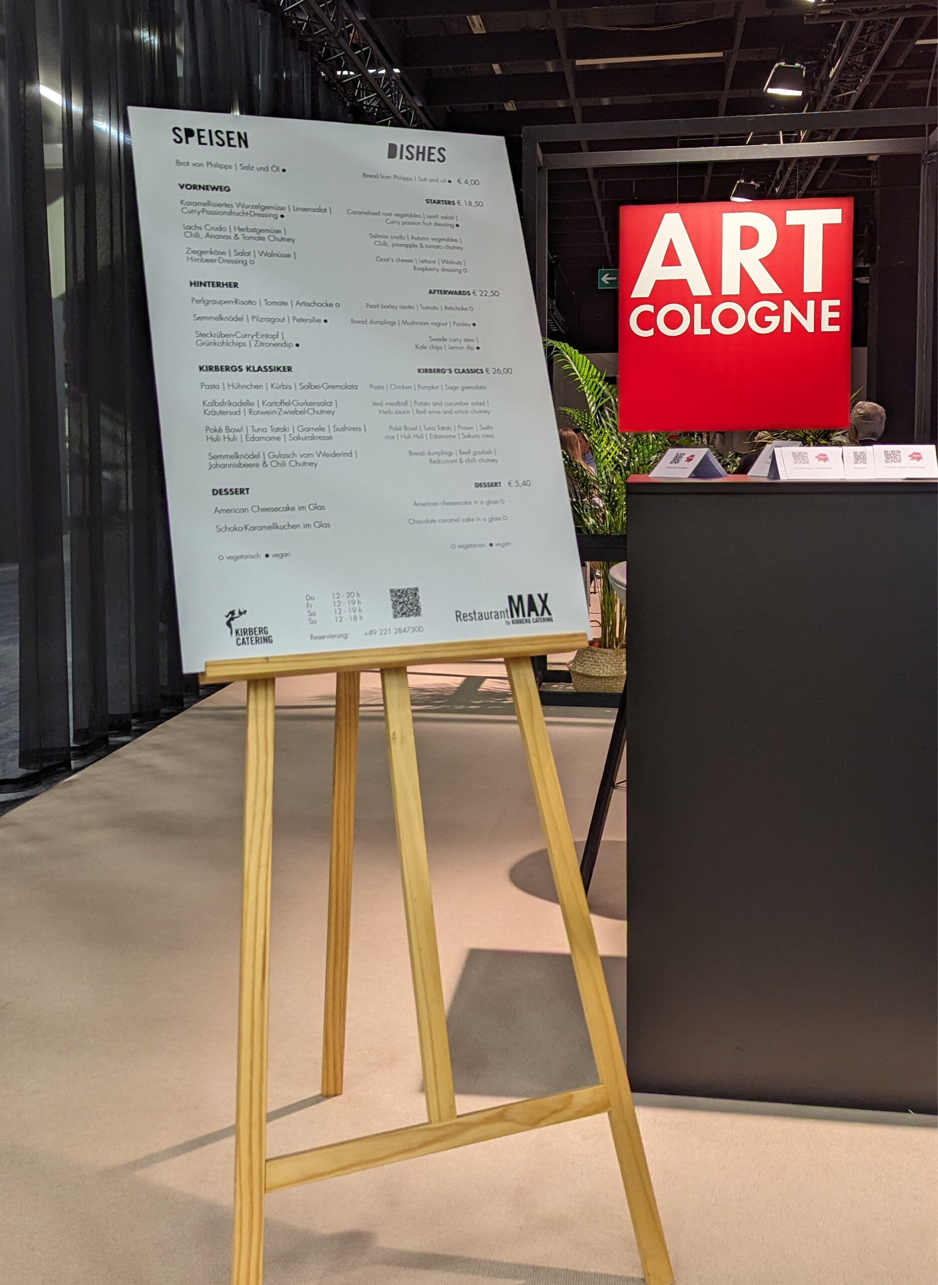 Eine Speisekarte auf einer Staffelei steht neben einem roten ART COLOGNE-Schild bei einer Indoor-Veranstaltung, die das Messecatering für Veranstaltungen präsentiert.
