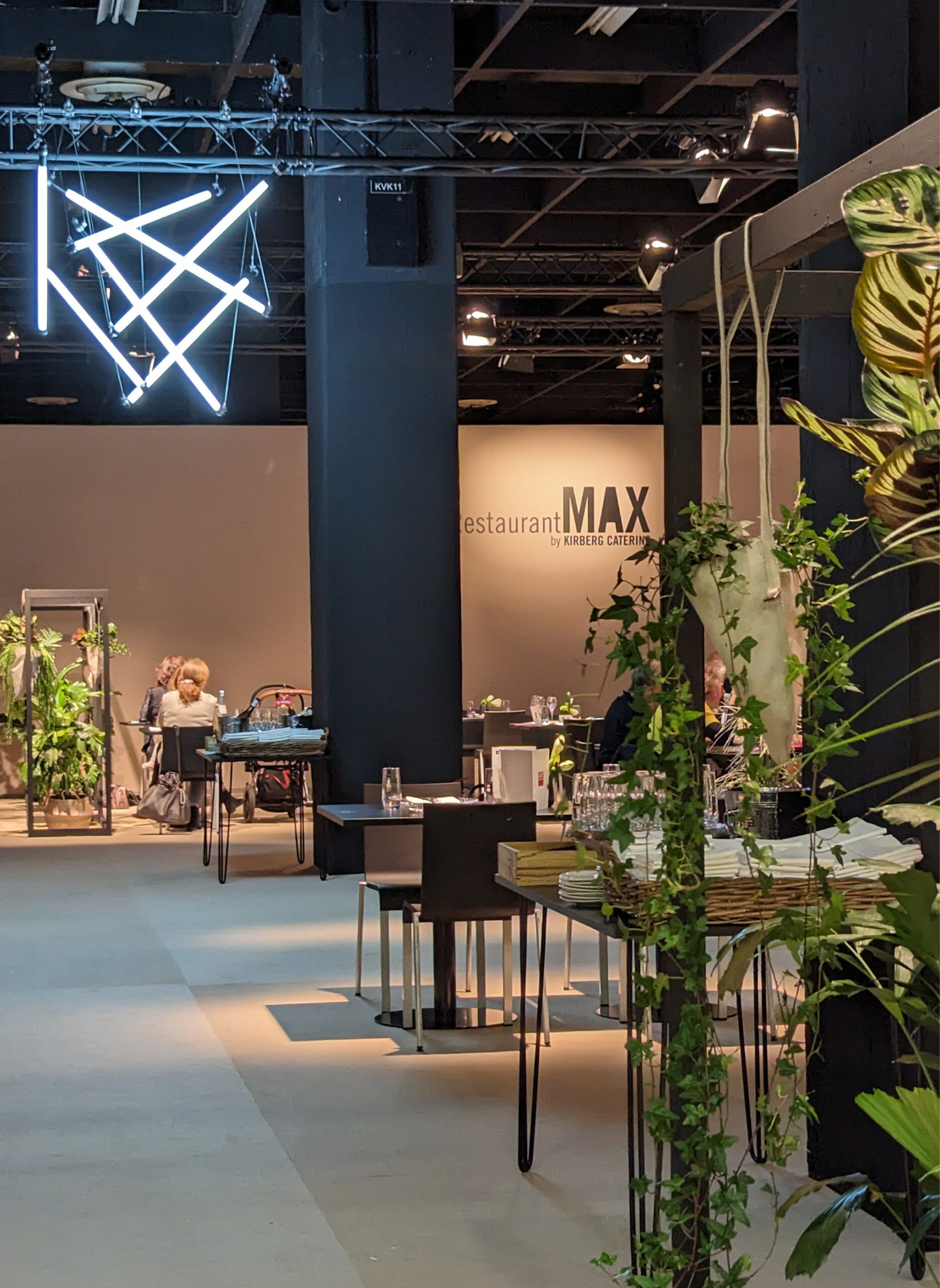 Modernes Restaurantinterieur mit geometrischen Leuchten, Grünpflanzen und Menschen, die im Hintergrund speisen - ideal für Messecatering für Veranstaltungen.