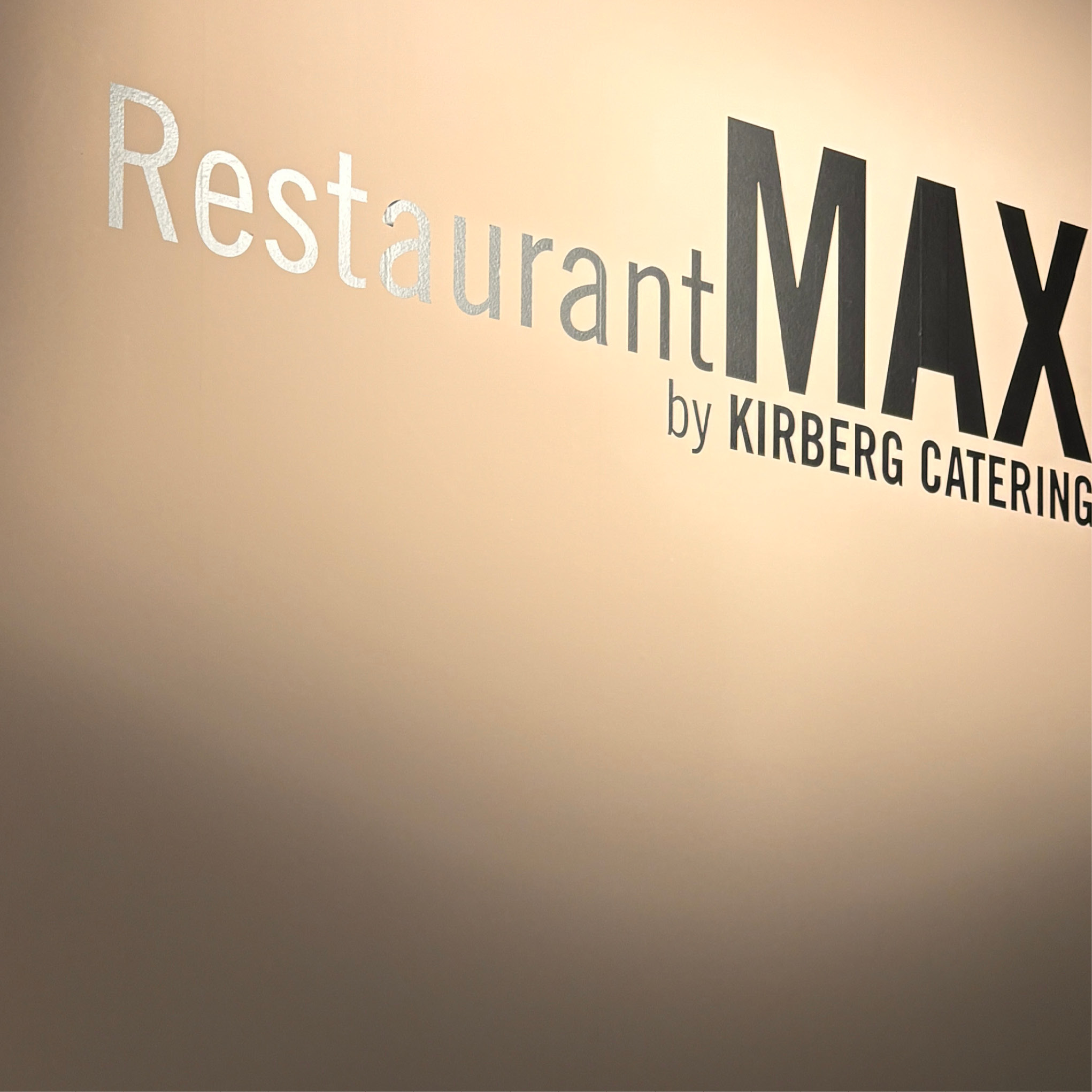 Auf dem Wandschild steht in großen schwarzen und silbernen Buchstaben auf beigem Hintergrund "Restaurant MAX by Kirberg Catering", das Messecatering für Veranstaltungen anbietet.