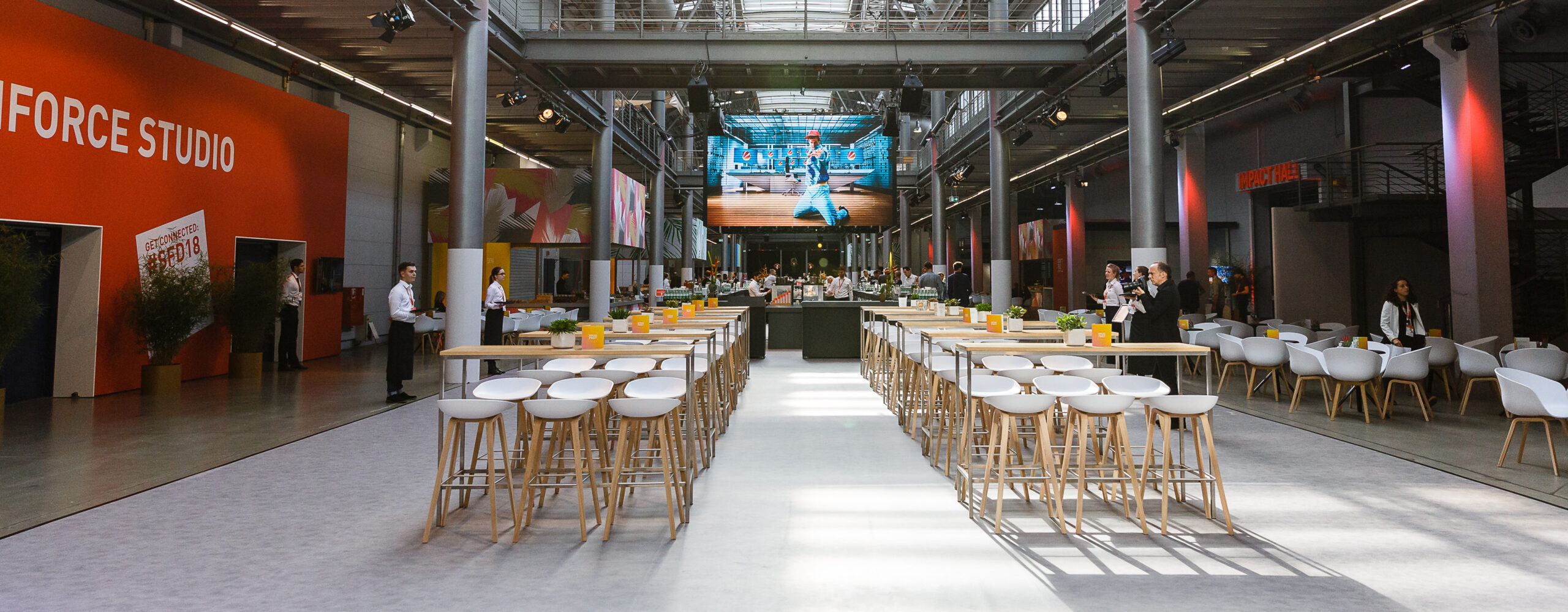 Moderner Veranstaltungsraum mit Stehtischen und Hockern, einer großen digitalen Leinwand im hinteren Teil des Raumes und Catering für Tagungen und Konferenzen auf Anfrage.