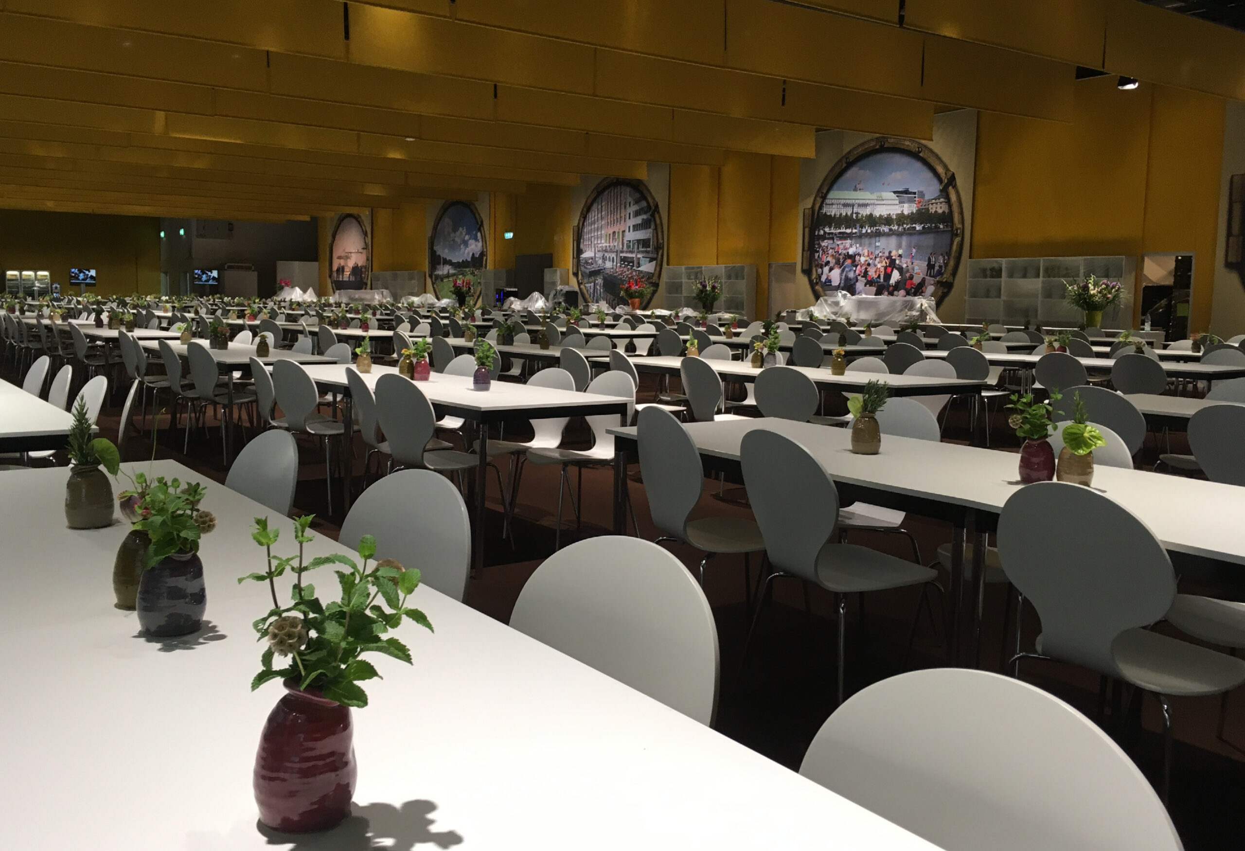 Große Cafeteria mit weißen Tischreihen, modernen Stühlen und kleinen Blumenvasen auf jedem Tisch - perfekt für Catering für Tagungen und Konferenzen.