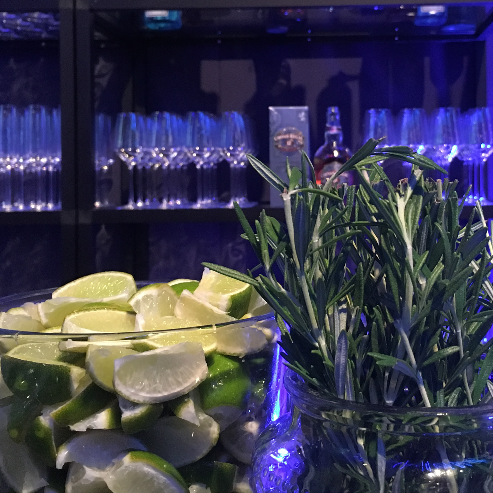 Glasbehälter mit Limettenspalten und Rosmarinzweigen vor Gläsern und Flaschen auf einem Barregal, ideal für hochwertiges Catering für Tagungen und Konferenzen.