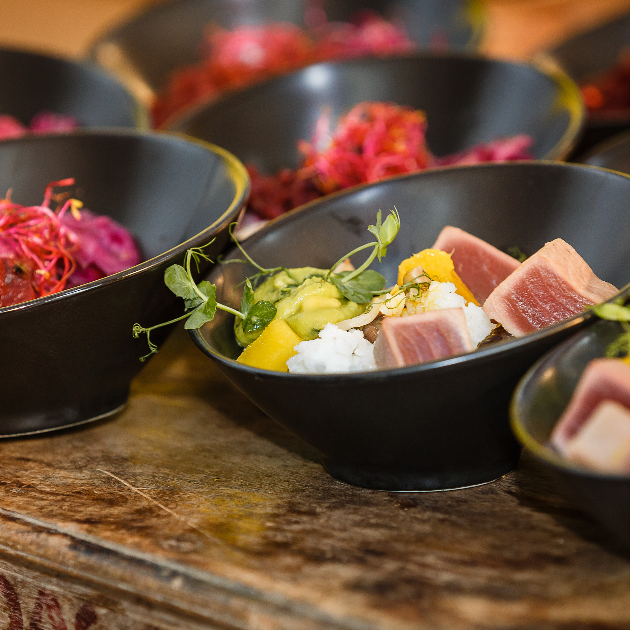 Schwarze Schalen mit gewürfeltem Thunfisch, Reis, Gemüse und Kräutern auf einer hölzernen Oberfläche, perfekt für Catering für Tagungen und Konferenzen, mit weiteren Schalen im Hintergrund.