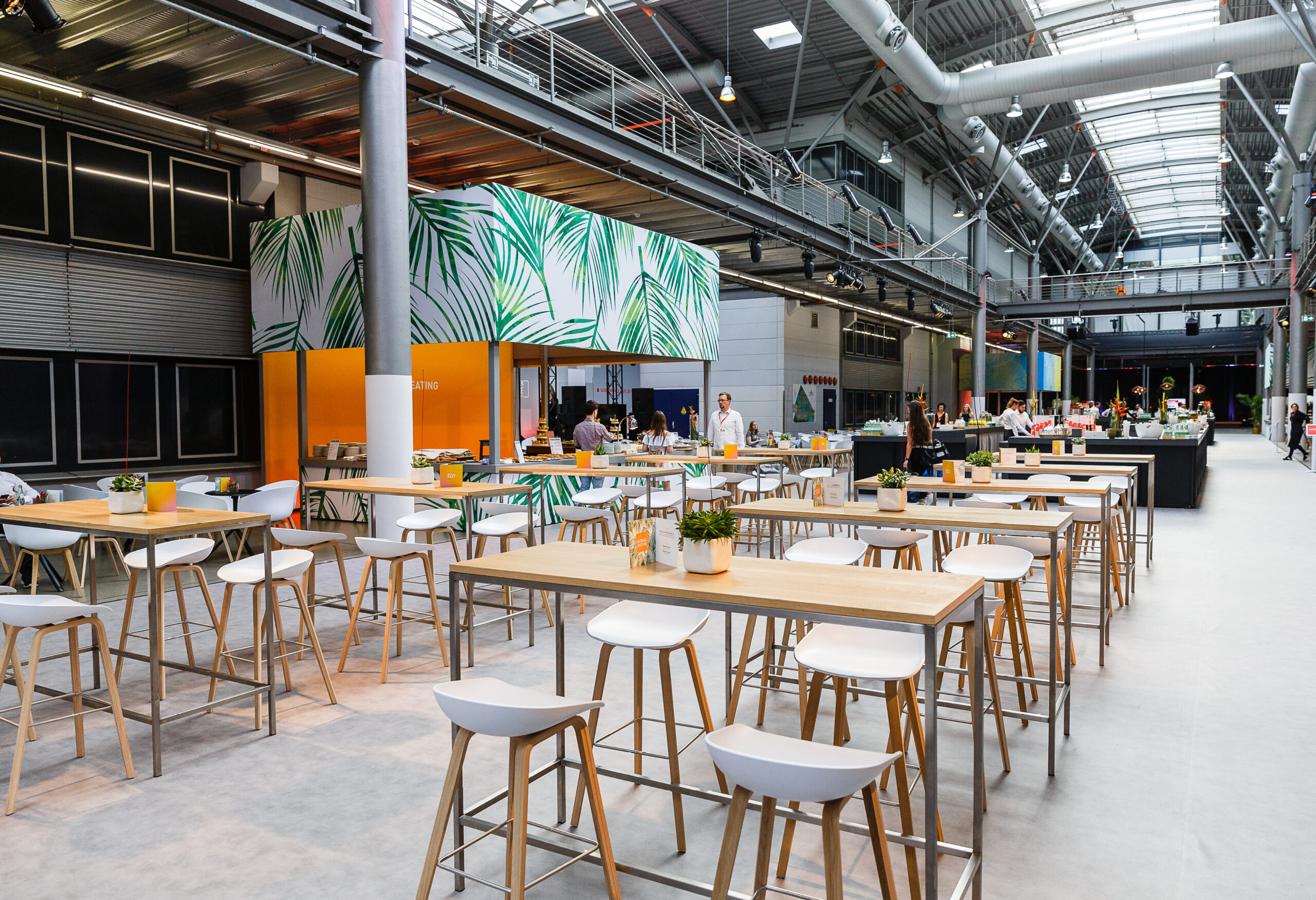 Geräumige, moderne Cafeteria mit hohen Tischen, weißen Stühlen und Topfpflanzen in einem Gebäude im Industriestil, ideal für Catering für Tagungen und Konferenzen.