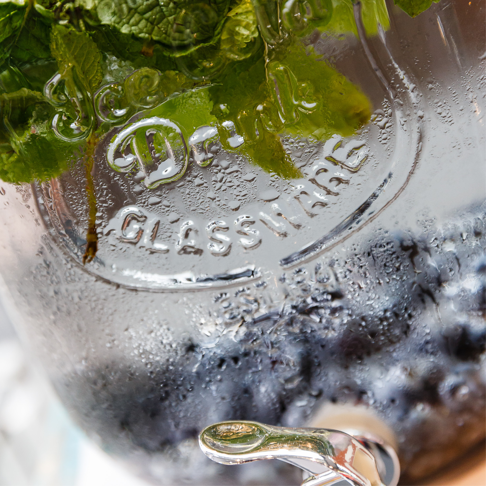 Nahaufnahme eines gläsernen Getränkespenders mit Kondenswasser, gefüllt mit Blaubeeren und Minzblättern - ideal für Catering für Tagungen und Konferenzen.