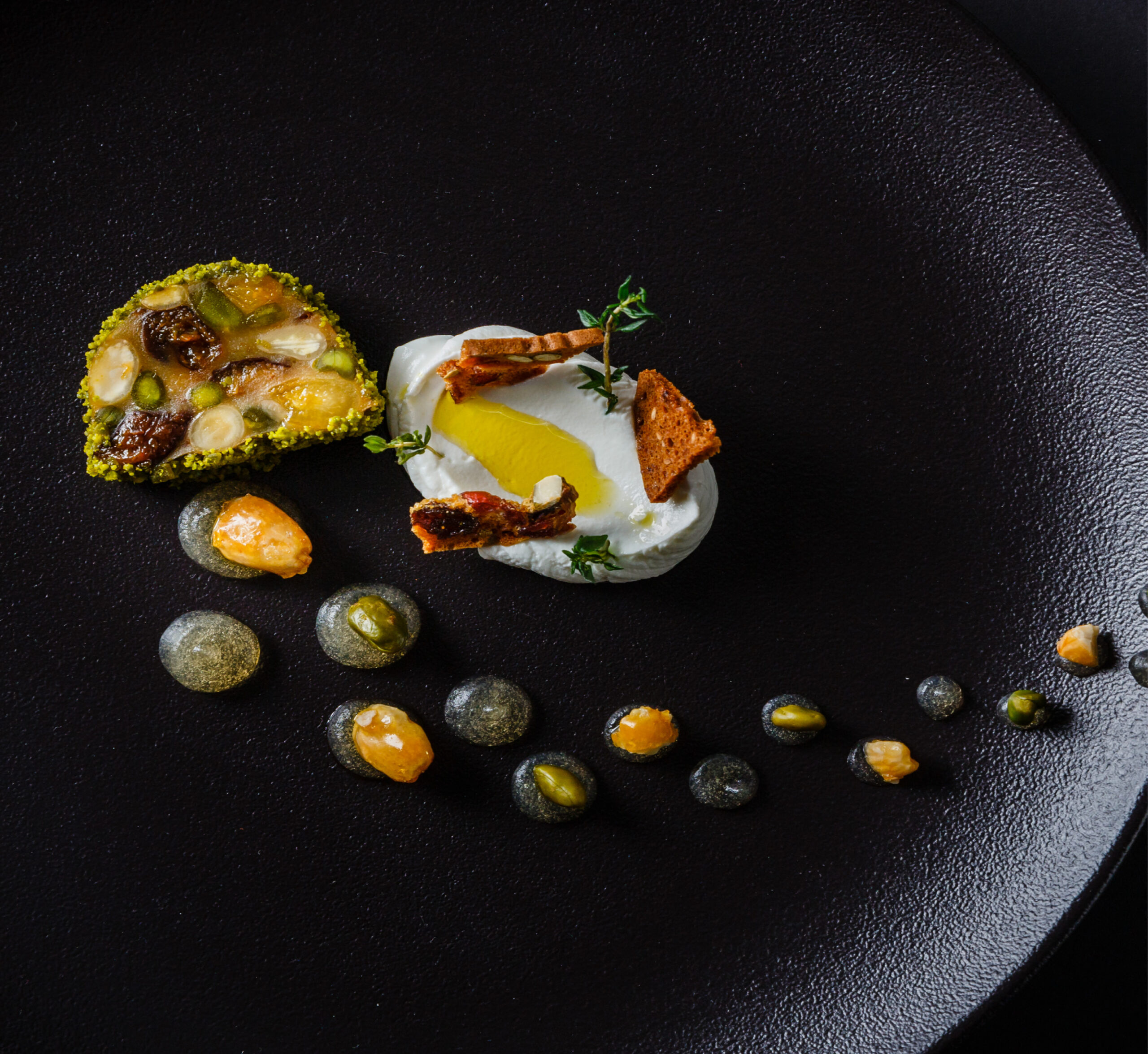 Modernes Tellergericht mit Ziegenkäse, Nüssen, Samen, Kräutern und Soßenpunkten auf einem schwarzen Teller.