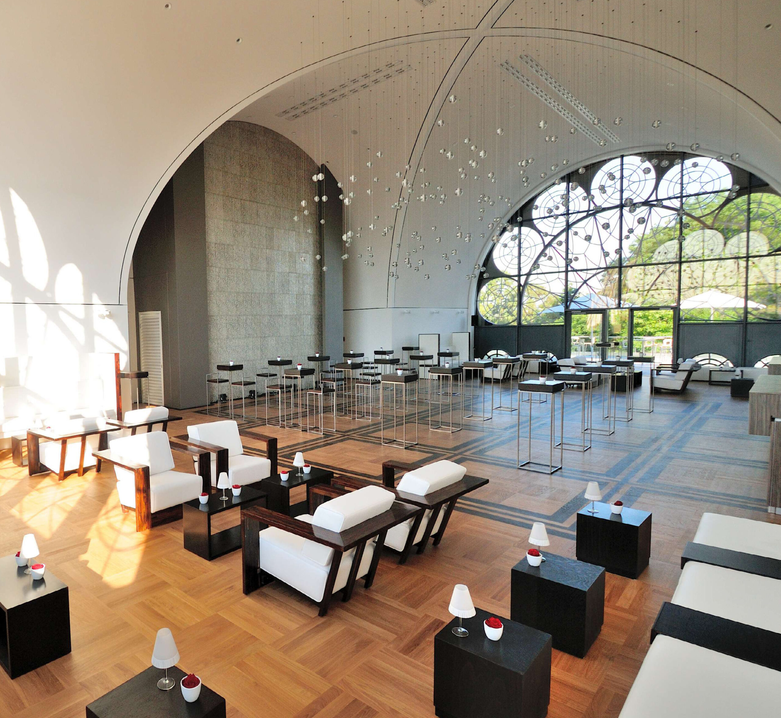 Geräumige, moderne Lounge mit weißen Sitzmöbeln, Holzböden und großen Bogenfenstern mit dekorativen Mustern - ideal für Catering für Tagungen und Konferenzen.