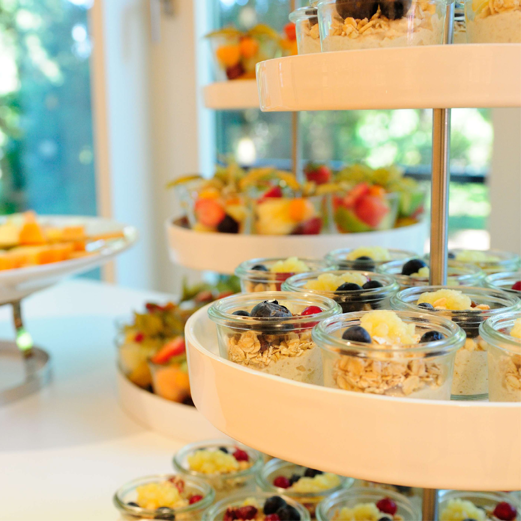 Kleine Gläschen mit Parfait, Müsli und Früchten sind auf einem hellen, modernen Buffet auf einem Stufentablett angeordnet - ideal für Catering für Tagungen und Konferenzen.