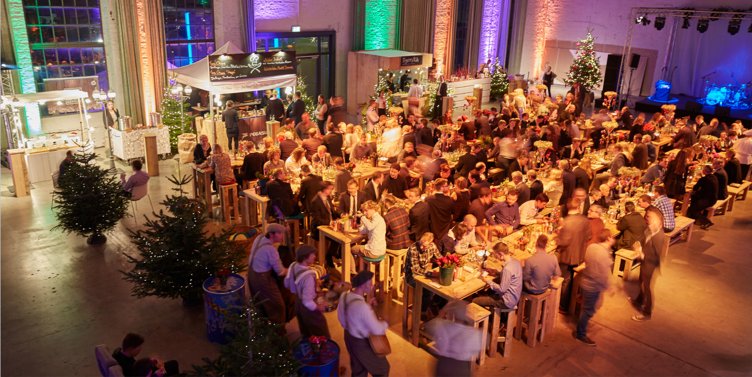 Ein festliches Indoor-Event mit langen Tischen, Weihnachtsbäumen, bunter Beleuchtung und exquisitem Weihnachtsfeiern Catering in Köln.