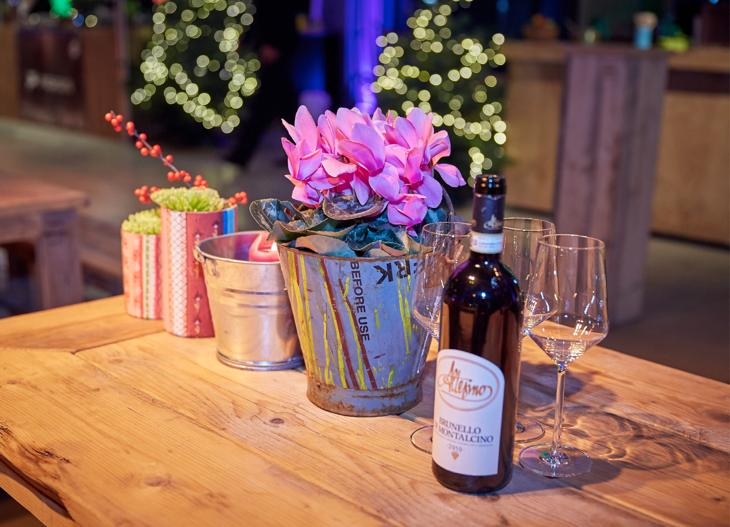 Eine Flasche Rotwein, zwei Gläser und rosa Blumen auf einem Holztisch mit festlicher Beleuchtung im Hintergrund - perfekt für das Weihnachtsfeiern Catering in Köln.