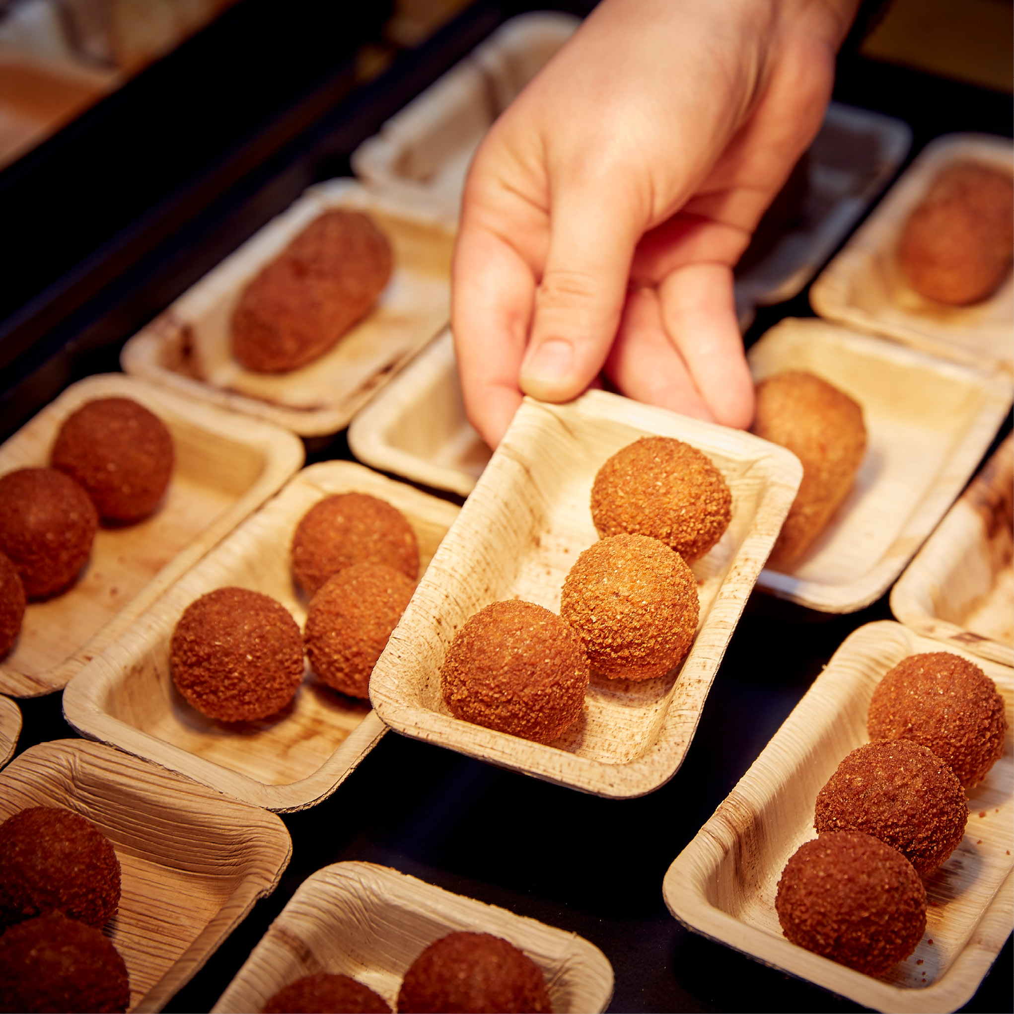 Eine Hand platziert ein Tablett mit runden, frittierten Snacks zwischen anderen identischen Tabletts auf einem Präsentationstisch - perfekt für das Weihnachtsfeiern Catering in Köln.