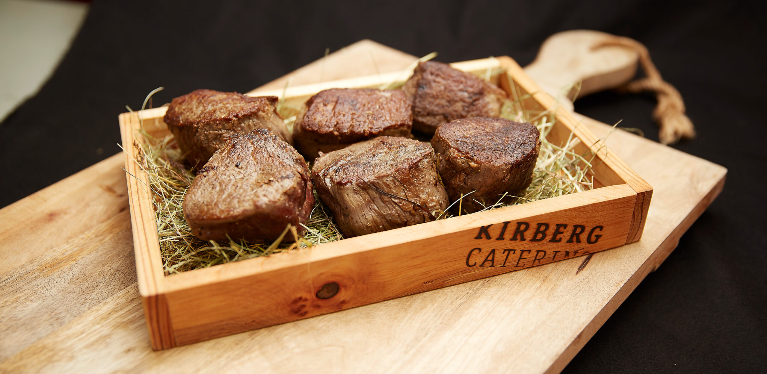 Sechs gekochte Rindersteaks auf Heu, serviert in einer Holzkiste mit der Aufschrift KIRBERG CATERING, auf einem Holzbrett - perfekt für das Weihnachtsfeiern Catering in Köln.