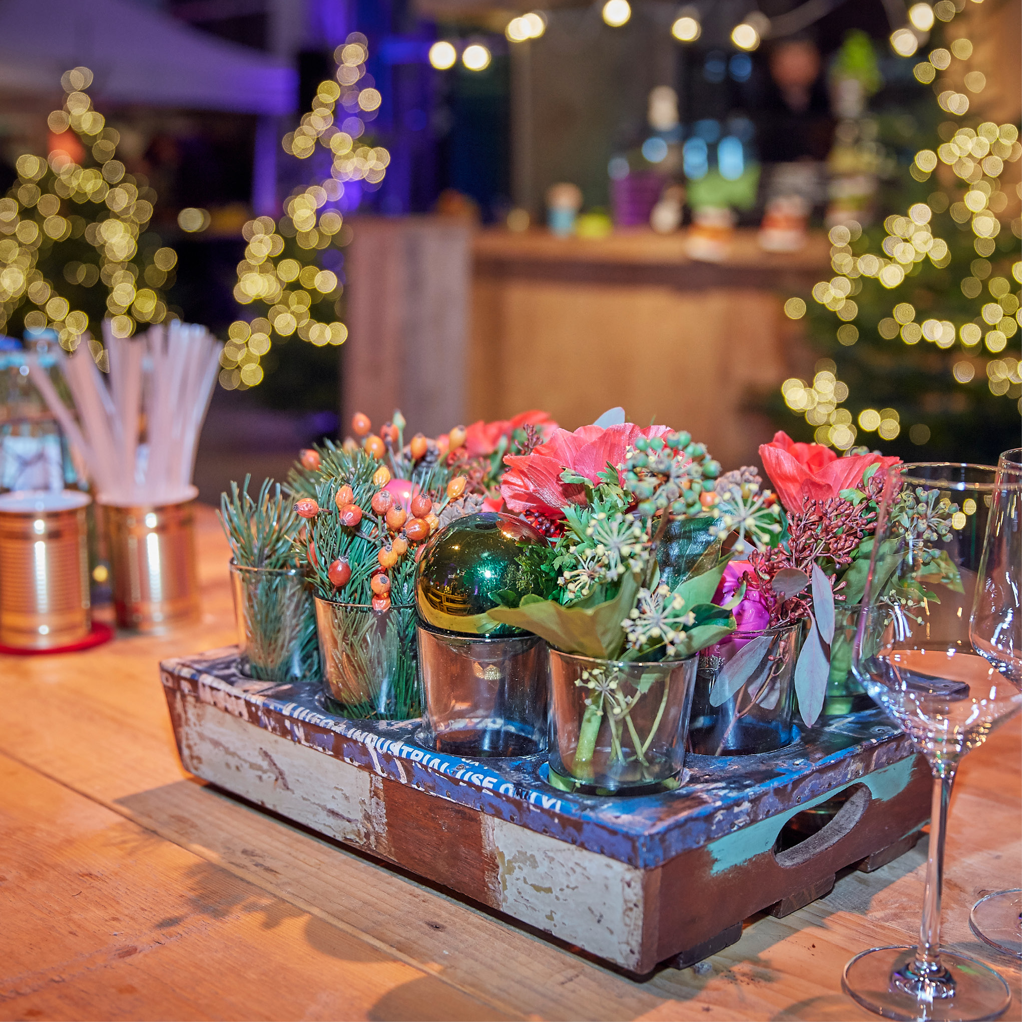 Ein Holztablett mit festlichen Blumen und Ornamenten auf einem Tisch, perfekt für das Weihnachtsfeiern Catering in Köln, mit Lichterketten und blinkenden Weihnachtsbäumen im Hintergrund.