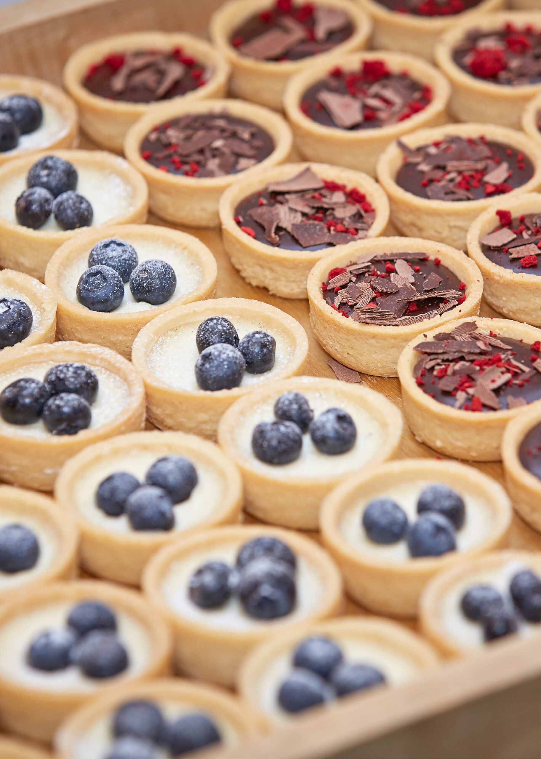 Verschiedene Mini-Törtchen mit Blaubeeren, Schokolade und roten Streuseln, aufgereiht auf einem Tablett - perfekt für das Weihnachtsfeiern Catering in Köln.