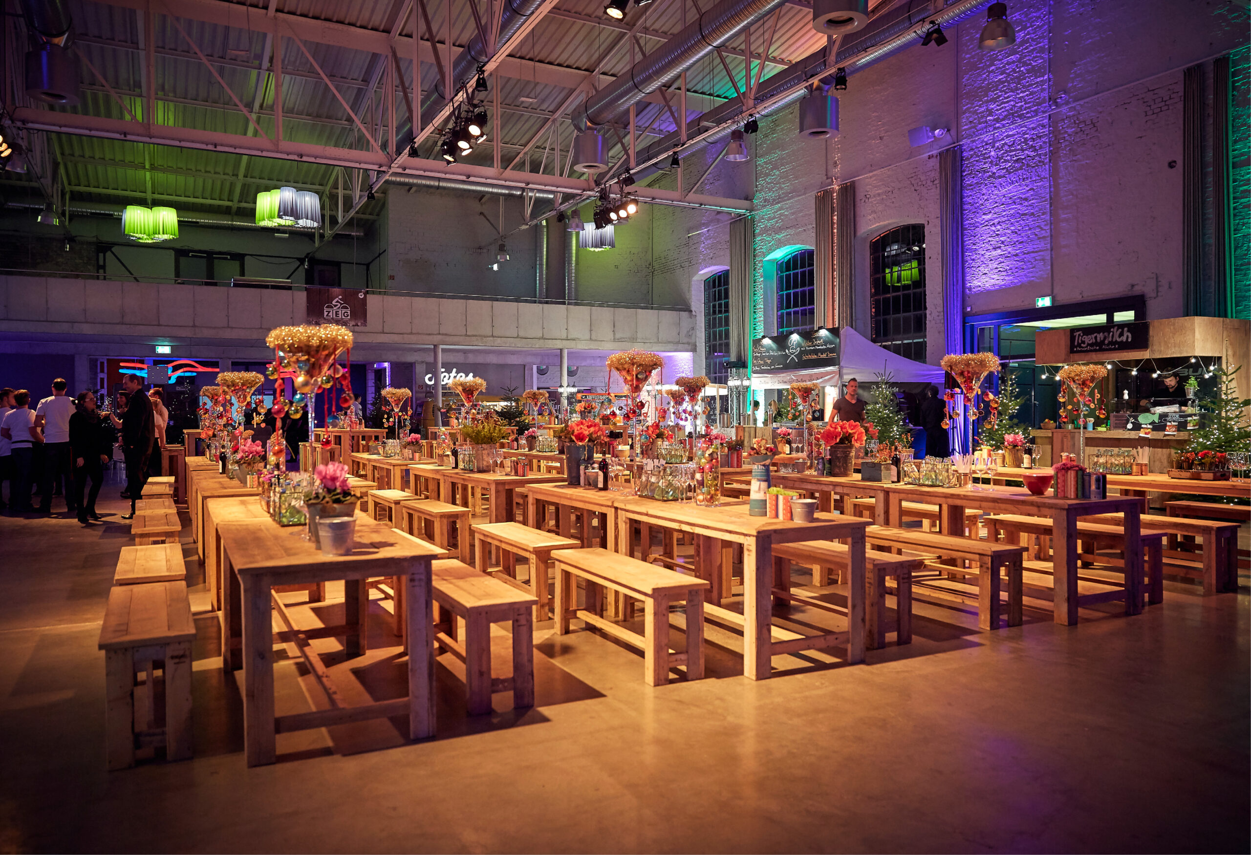 Geräumige Veranstaltungshalle mit Holztischen, Bänken, farbenfrohen Blumenkränzen und stimmungsvoller Beleuchtung - perfekt für Weihnachtsfeiern Catering in Köln.