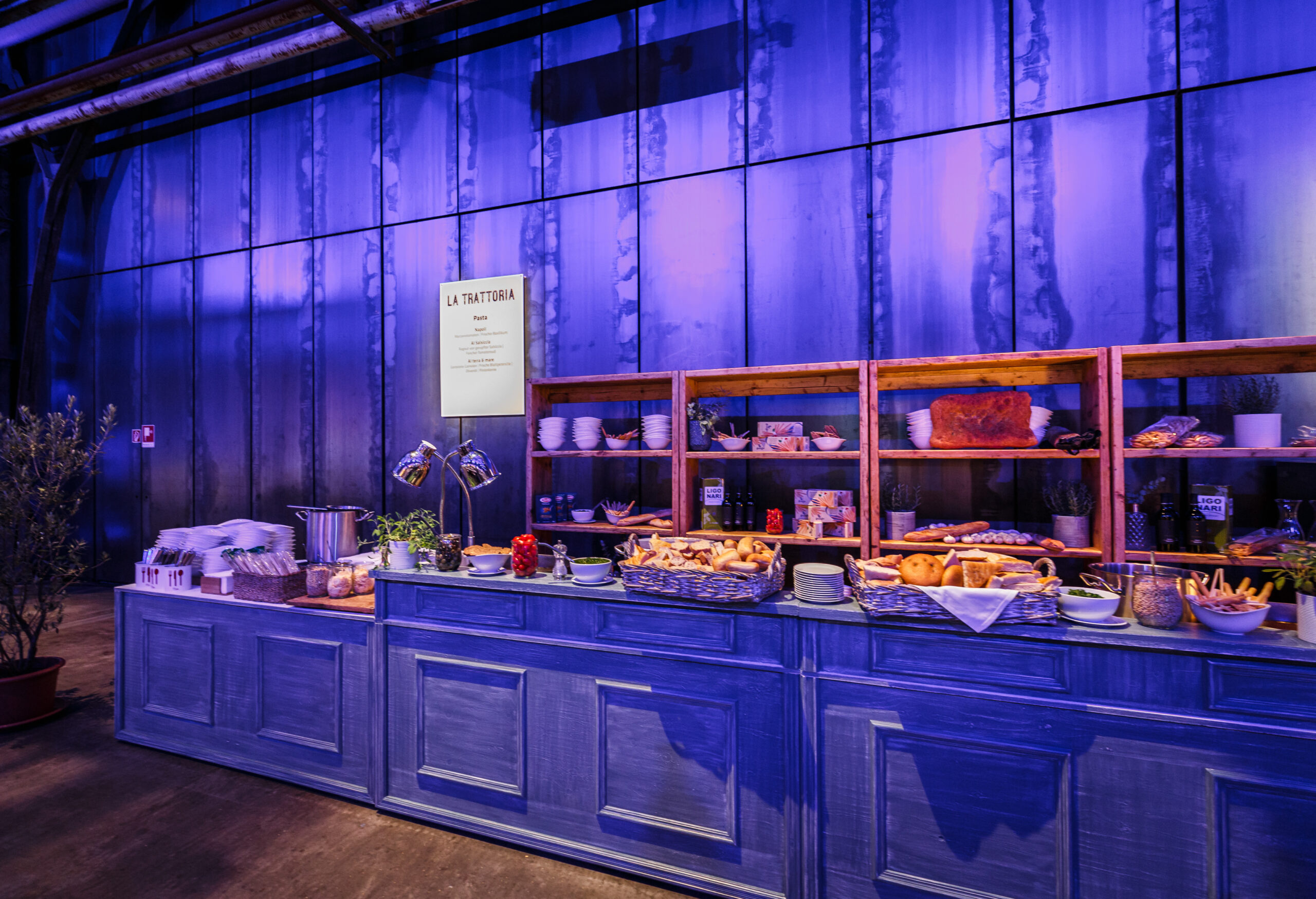 Buffet-Tisch mit verschiedenen Speisen, Tellern und Utensilien vor einer blau beleuchteten Wand in einem Industrieraum - ideal für Weihnachtsfeiern Catering in Köln.