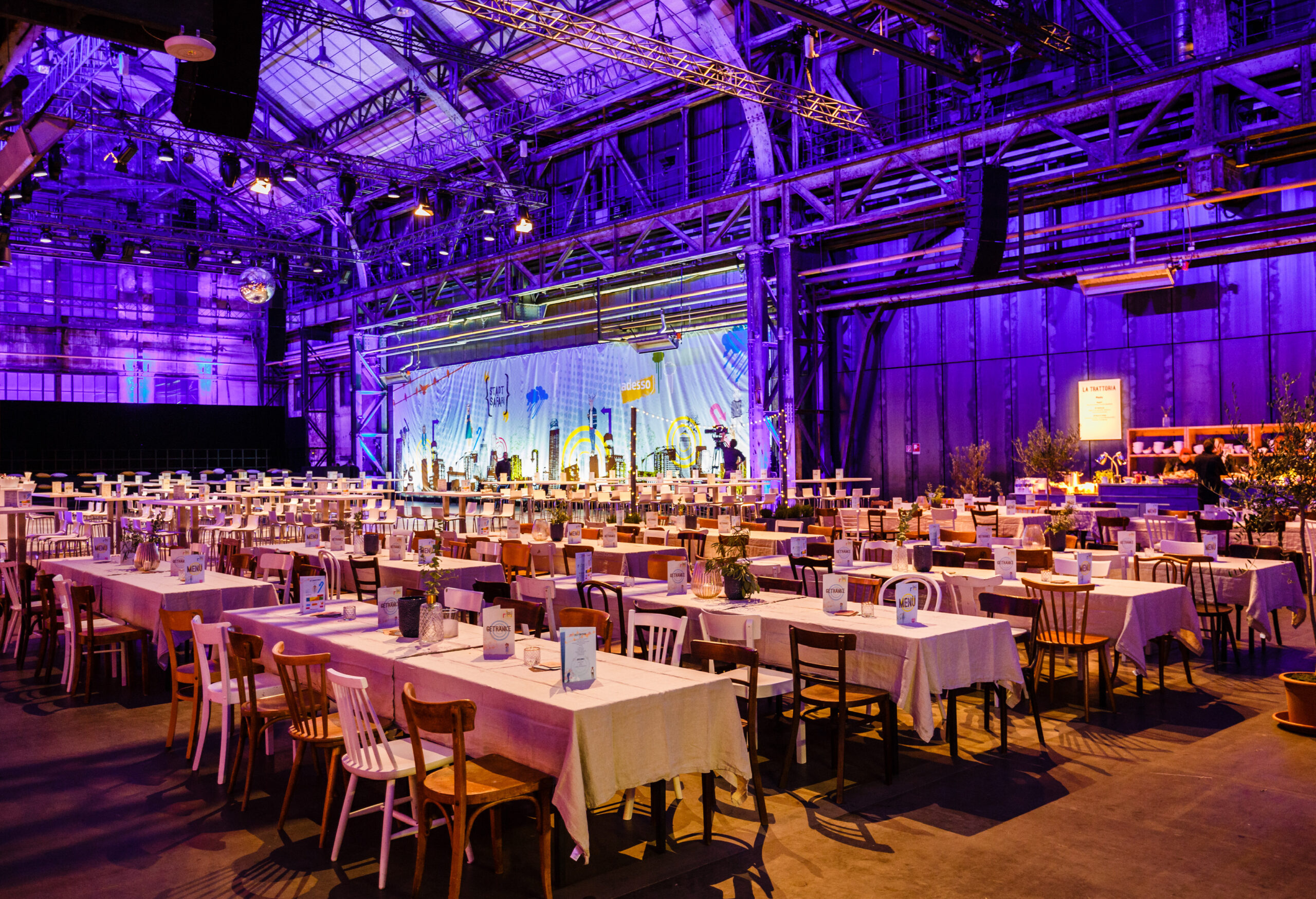 Ein großer Veranstaltungssaal mit Tisch- und Stuhlreihen, dekoriert mit lila Beleuchtung und einer Bühne im Hintergrund - perfekt für Weihnachtsfeiern Catering in Köln.