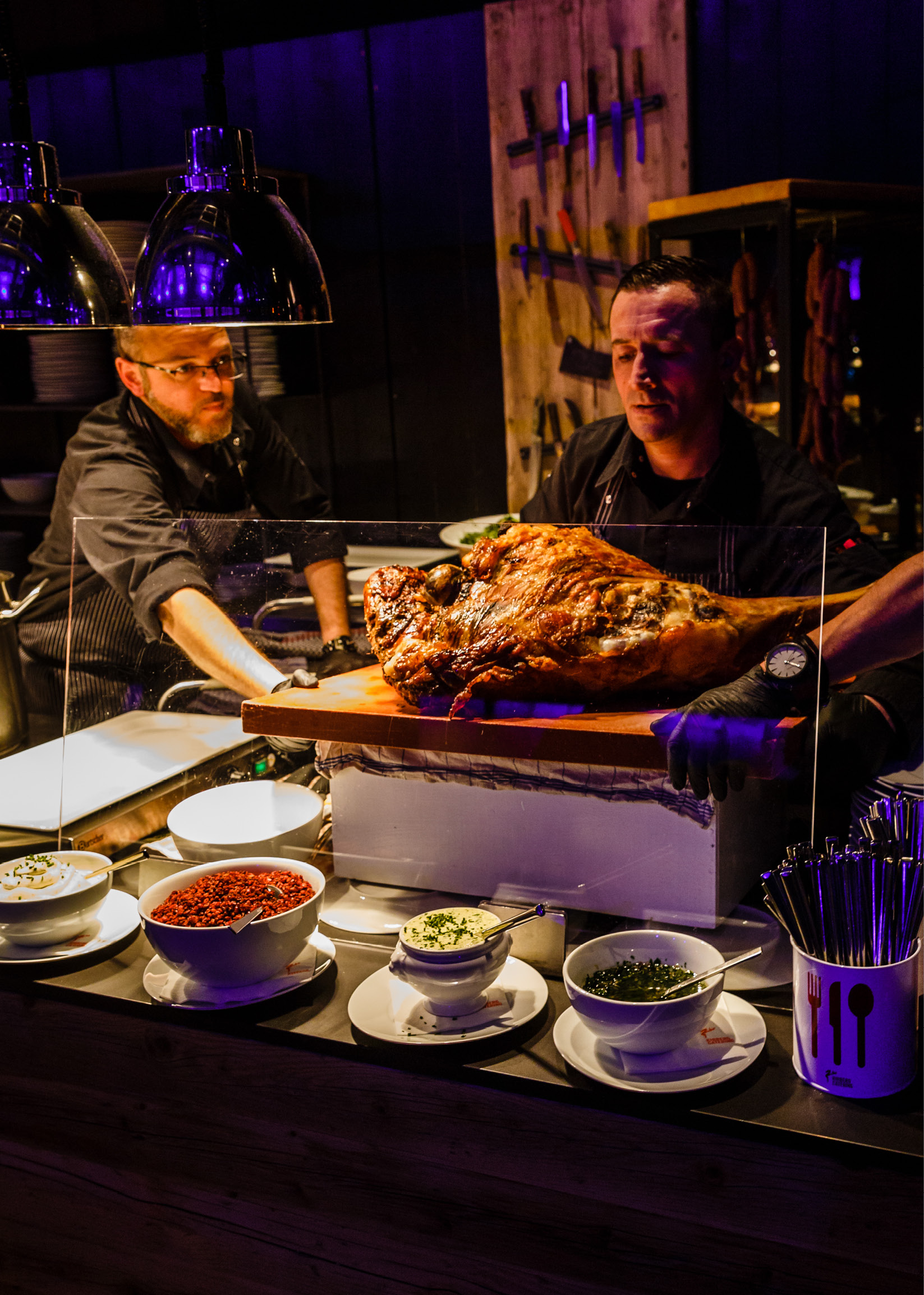 Zwei Köche präsentieren ein großes gebratenes Fleisch auf einem Brett, umgeben von Schalen mit Gewürzen und Utensilien.