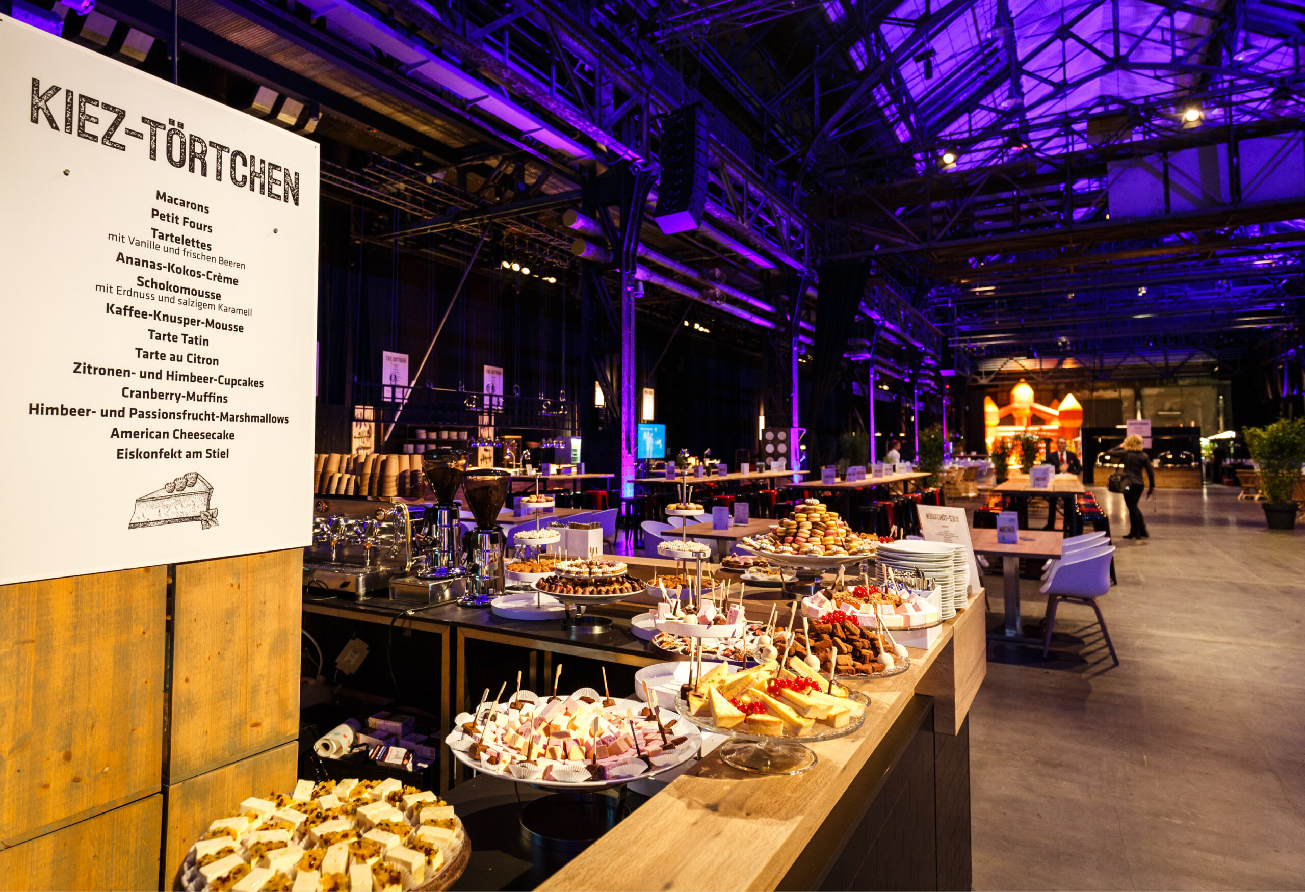 Genießen Sie ein Dessertbuffet mit verschiedenen Kuchen und Torten, perfekt für das Weihnachtsfeiern Catering in Köln, in einem großzügigen, lila beleuchteten Veranstaltungsraum mit Industriecharakter.