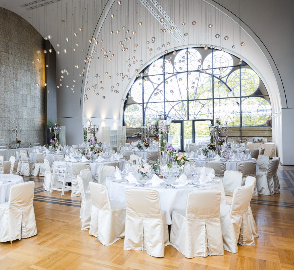Eleganter Bankettsaal mit runden Tischen, weißen Stühlen, Blumenschmuck und großen Bogenfenstern - perfekt für alle, die Eventlocations Köln für einen besonderen Anlass mieten möchten.