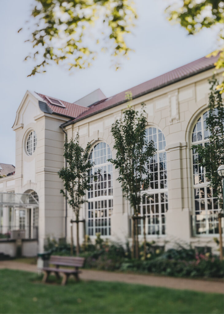 Ein cremefarbenes Gebäude mit gewölbten Fenstern, Bäumen und einer Bank auf einer Wiese - perfekt, wenn Sie auf der Suche nach Eventlocations Köln mieten für Ihre nächste Zusammenkunft sind.