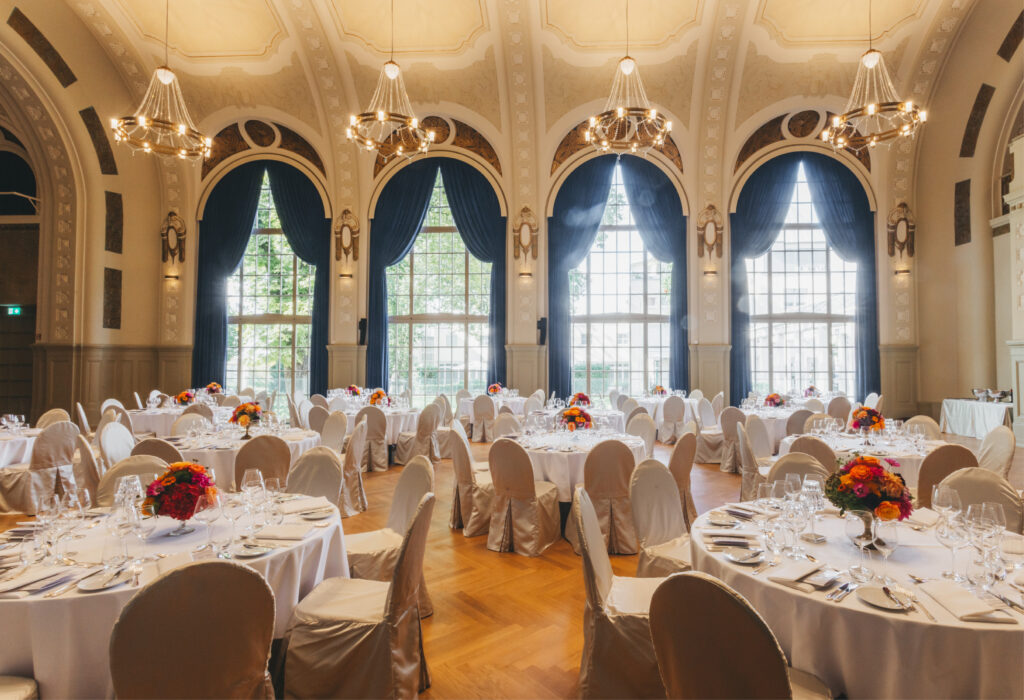 Eleganter Bankettsaal mit runden Tischen, weißen Tischdecken, Blumenschmuck und großen Bogenfenstern.