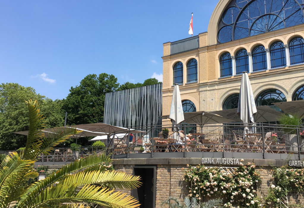 Außencafé mit Sonnenschirmen und Menschen, die an einem sonnigen Tag auf einer Terrasse neben einem großen Gebäude sitzen - perfekt für Versammlungen oder Feiern.