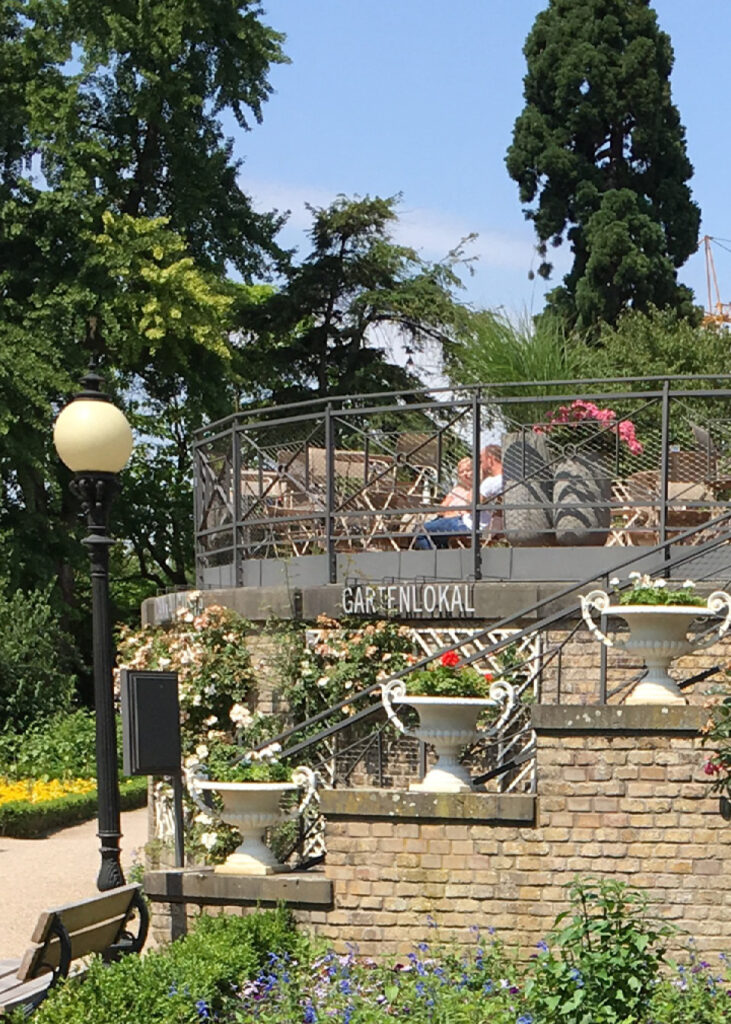 Erhöhte Außenterrasse mit Sitzgelegenheiten, umgeben von Grün und einem Gartenweg unterhalb - perfekt für alle, die Eventlocations Köln in einer schönen, ruhigen Umgebung mieten möchten.