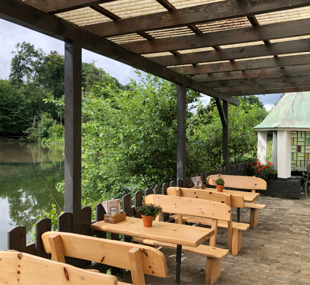 Ein hölzerner Sitzbereich im Freien am See, unter einer Pergola, mit Bäumen und einem kleinen Gebäude im Hintergrund.
