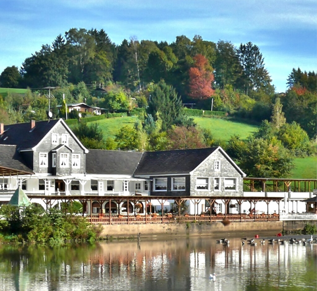 Ein großes Landhaus mit Veranda liegt an einem ruhigen See, umgeben von grünen Bäumen und Hügeln - ideal für diejenigen, die Eventlocations Köln mieten in einer ruhigen Umgebung suchen.