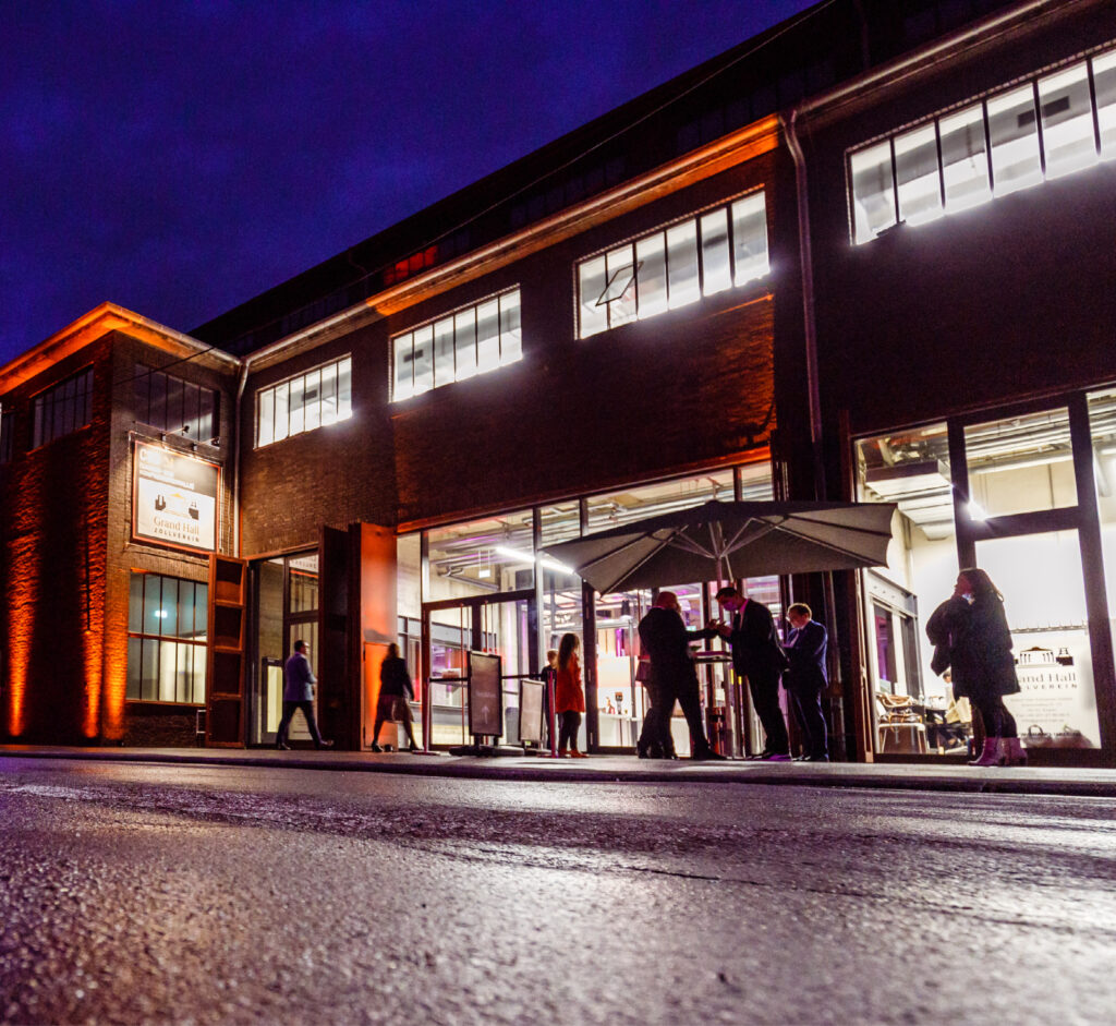 Vor einem hell erleuchteten Backsteingebäude stehen und gehen die Menschen bei Nacht, orangefarbene Lichter erhellen das Äußere - eine einladende Szene für diejenigen, die Eventlocations Köln mieten für besondere Veranstaltungen.