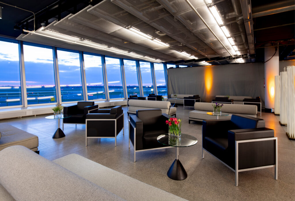 Moderne Flughafenlounge mit großen Fenstern, stilvollen Sitzgelegenheiten, Glastischen und Blumenarrangements bei Sonnenuntergang.