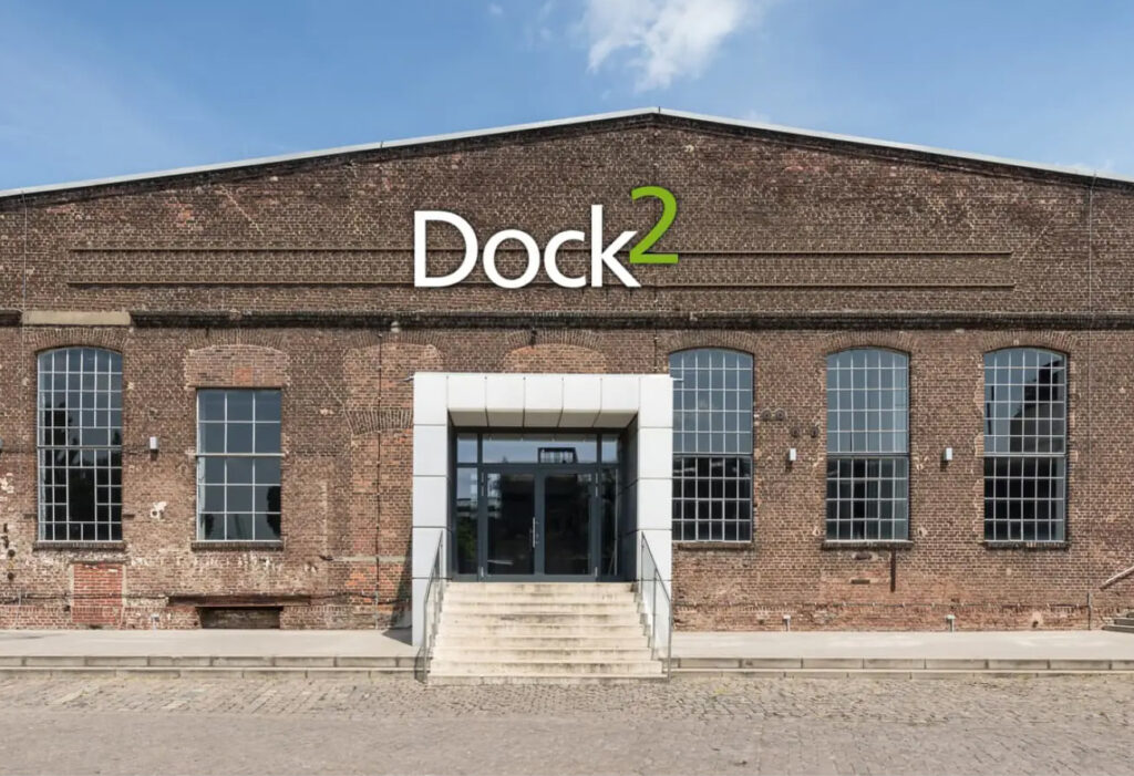 Frontansicht eines Backsteingebäudes mit der Bezeichnung Dock2 mit großen Fenstern und einem zentralen Eingang.