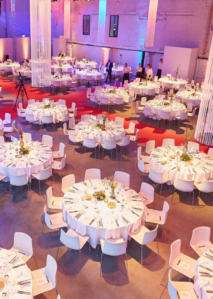 Ein großer Bankettsaal mit runden Tischen, die für eine Veranstaltung gedeckt sind, weißen Tischdecken und geordneten Stühlen - ideal für diejenigen, die Eventlocations Köln mieten möchten.