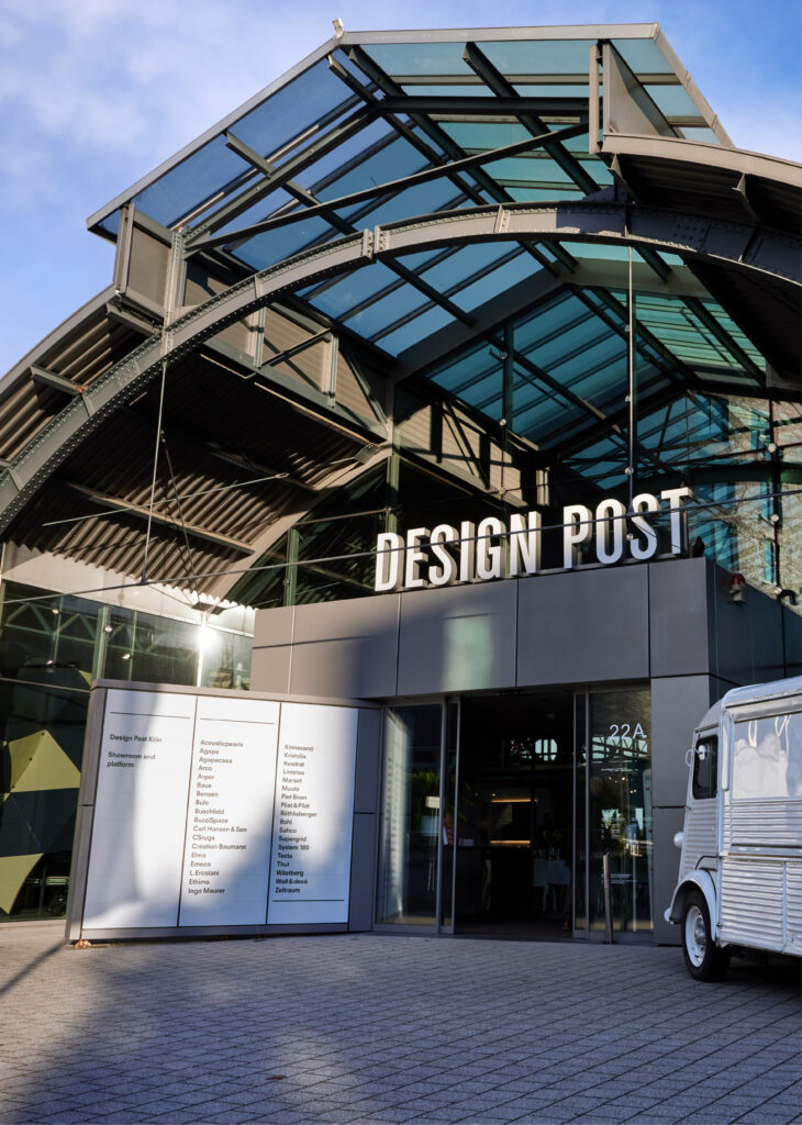 Ein gläserner Gebäudeeingang mit der Aufschrift DESIGN POST und ein klassischer weißer Lieferwagen, der vor dem Gebäude geparkt ist - die perfekte Inspiration, wenn Sie Eventlocations Köln mieten möchten.