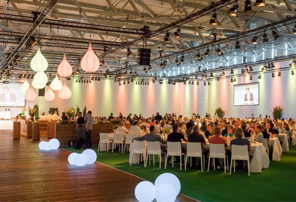 Großer Bankettsaal mit runden Tischen, sitzenden Gästen und bunten Hängelampen während einer formellen Veranstaltung.
