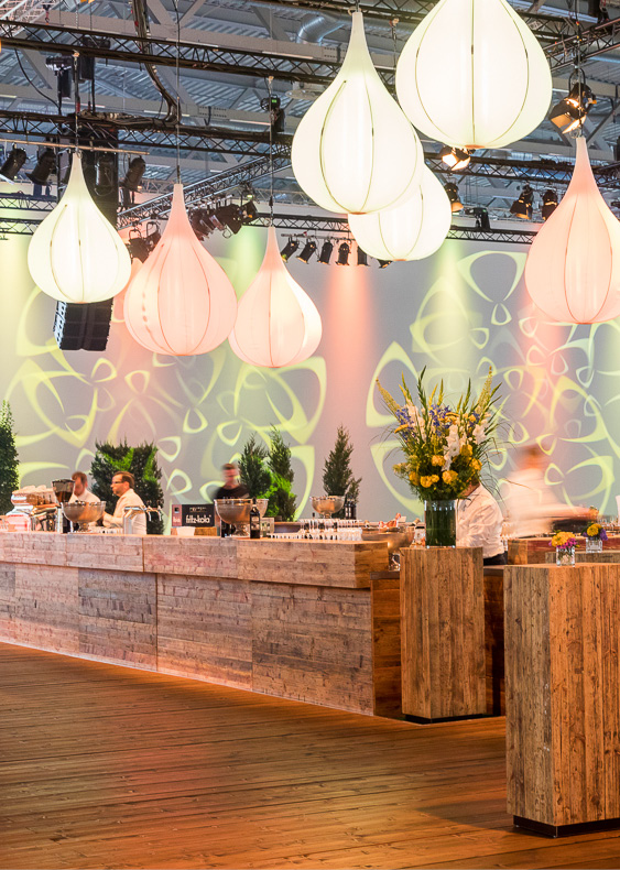 Eine moderne Eventbar mit Holztresen, hängenden weißen Lichtern und Köchen, die hinter dem Tresen Speisen zubereiten.