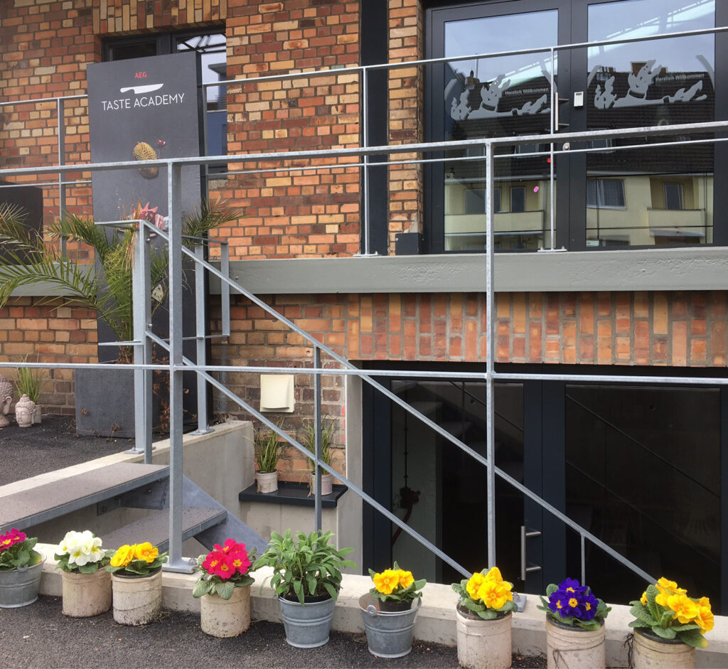 Topfblumen säumen eine Rampe vor einem Backsteingebäude mit einem Schild, auf dem "Taste Academy" steht, eine der einzigartigen Eventlocations, die Köln für besondere Anlässe mietet.