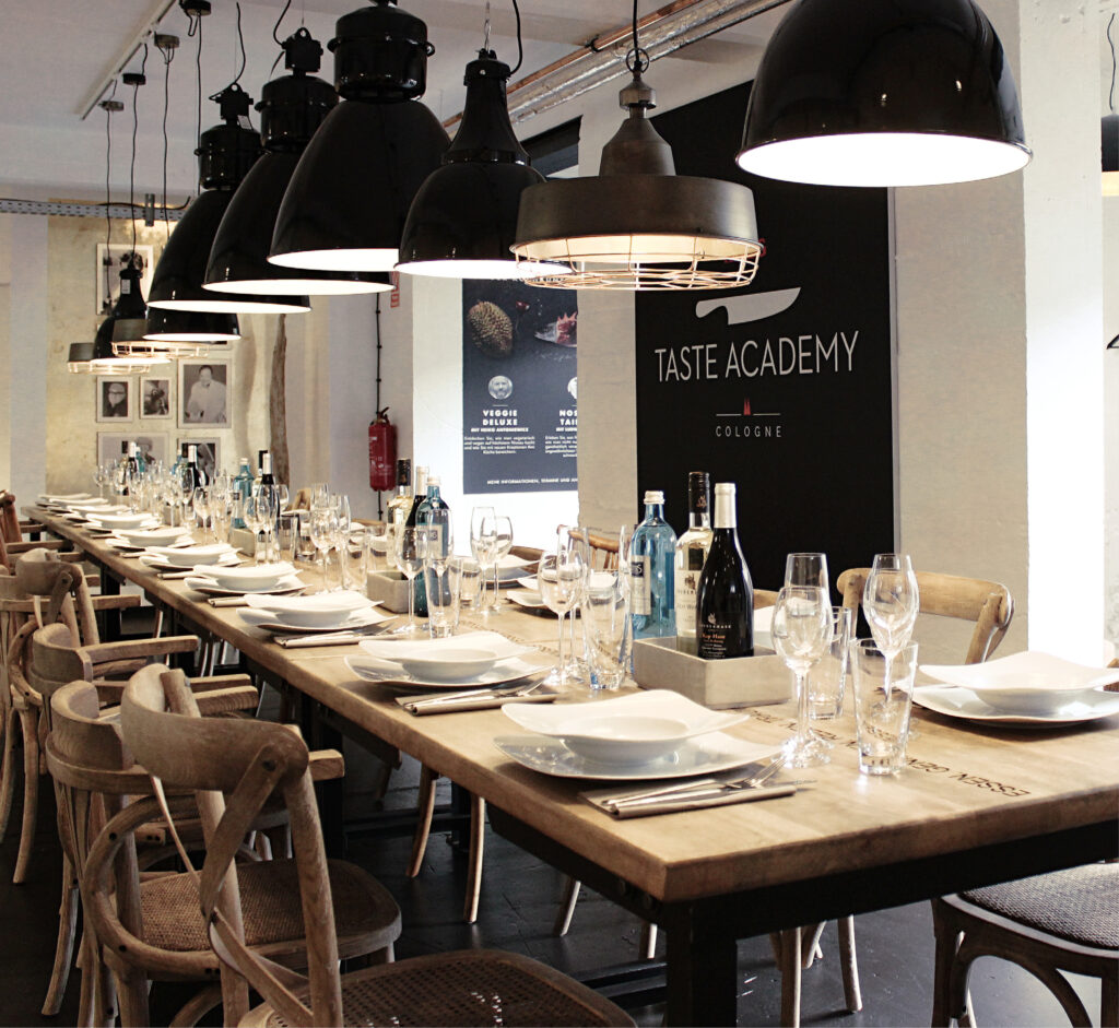 Ein langer Holztisch mit Tellern, Gläsern und Flaschen unter schwarzen Hängelampen in einem modernen Raum - die perfekte Inspiration für alle, die Eventlocations Köln mieten möchten.