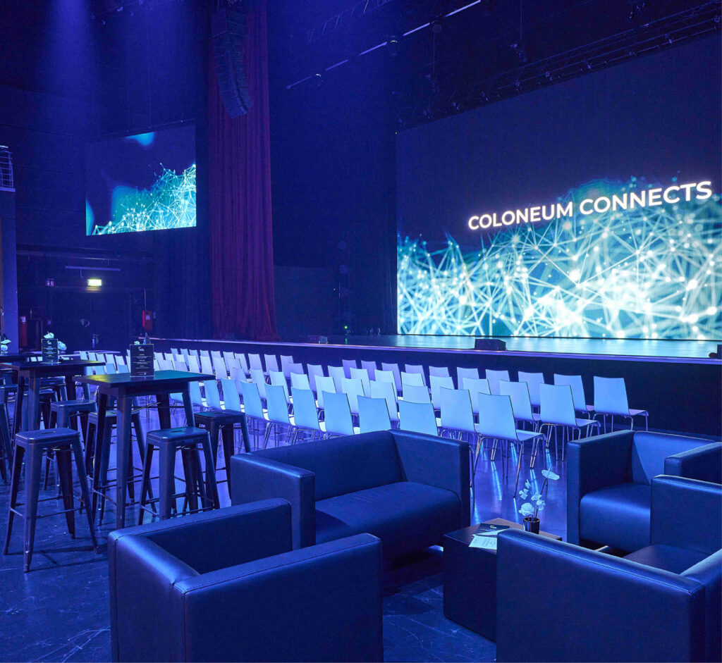 Moderner Veranstaltungsraum mit Stuhlreihen, Loungesitzen und einem großen Bildschirm mit Coloneum Connects - perfekt, wenn Sie Eventlocations Köln mieten möchten.