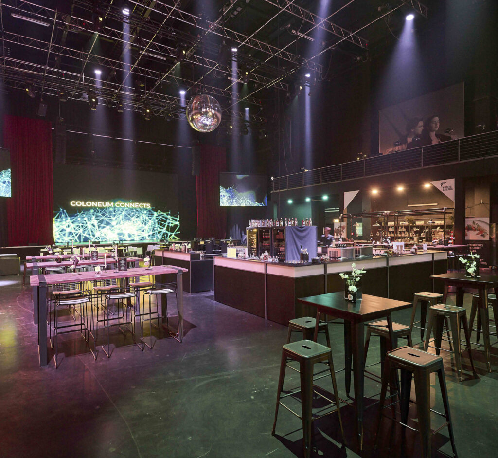 Großzügiger, moderner Veranstaltungsraum mit Stehtischen, Hockern, Bühne, Barbereich und großer Videoleinwand - ideal für alle, die Eventlocations Köln mieten wollen, für jeden Anlass.