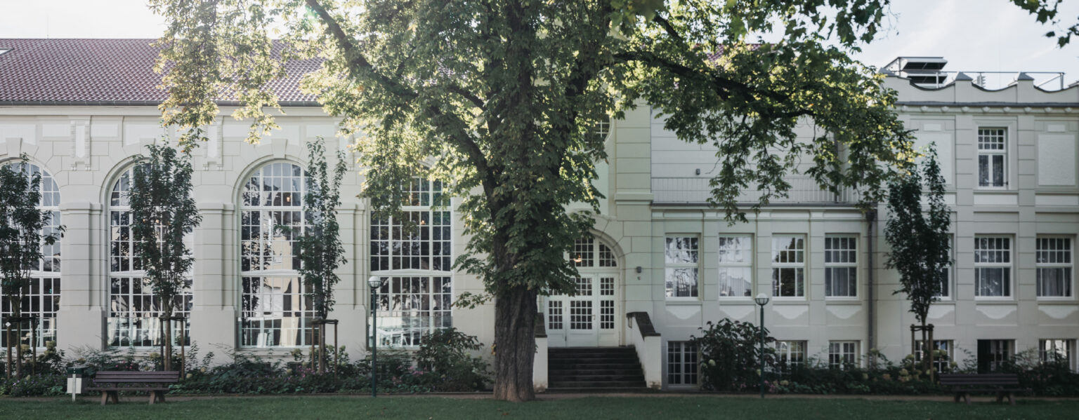 Großes weißes Gebäude mit hohen Fenstern, umgeben von Bäumen und Bänken, mit Blick auf eine Wiese - ein idealer Ort für Veranstaltungen oder Catering in Bad Honnef.