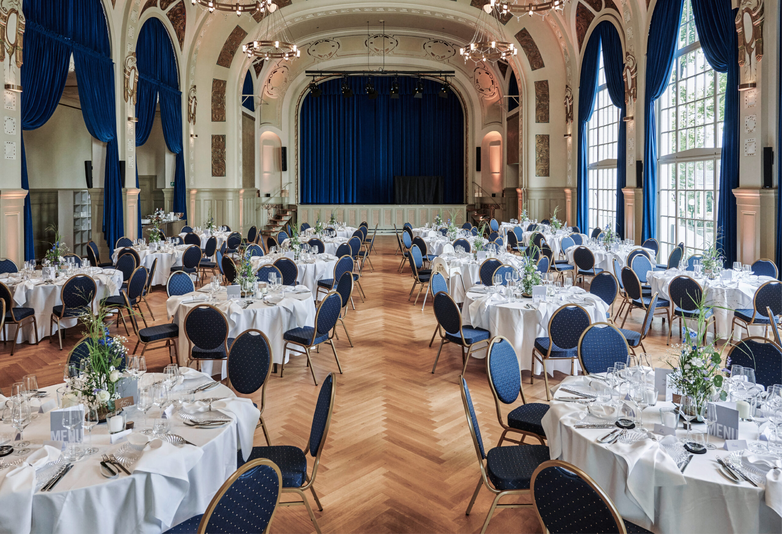 Eleganter Festsaal mit runden Tischen, die für eine Veranstaltung gedeckt sind, blauen Stühlen und großen Fenstern, die viel Tageslicht hereinlassen - perfekt für Veranstaltungen mit erstklassigem Catering in Bad Honnef.