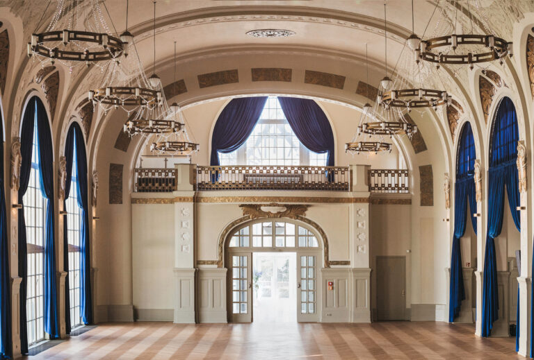 Eleganter Ballsaal mit hohen Bogenfenstern, blauen Vorhängen, Kronleuchtern und verschnörkelten architektonischen Details in Bad Honnef.