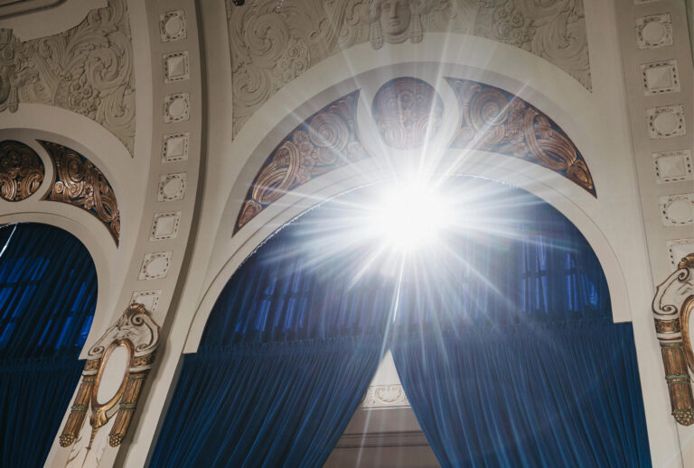 Sonnenlicht fällt durch kunstvolle Bogenfenster mit blauen Vorhängen und dekorativen weißen und goldenen Verzierungen und schafft eine einladende Atmosphäre, die sich perfekt für die Gastronomie in Bad Honnef eignet.