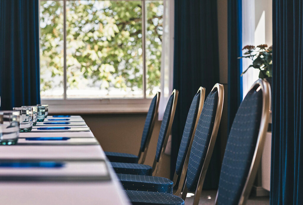 Leere blaue Stühle an einem Besprechungstisch mit Notizblöcken und Gläsern, in der Nähe eines sonnigen Fensters mit einer Pflanze – ein ideales Setup für Geschäftsgespräche oder Veranstaltungen mit Catering in Bad Honnef.