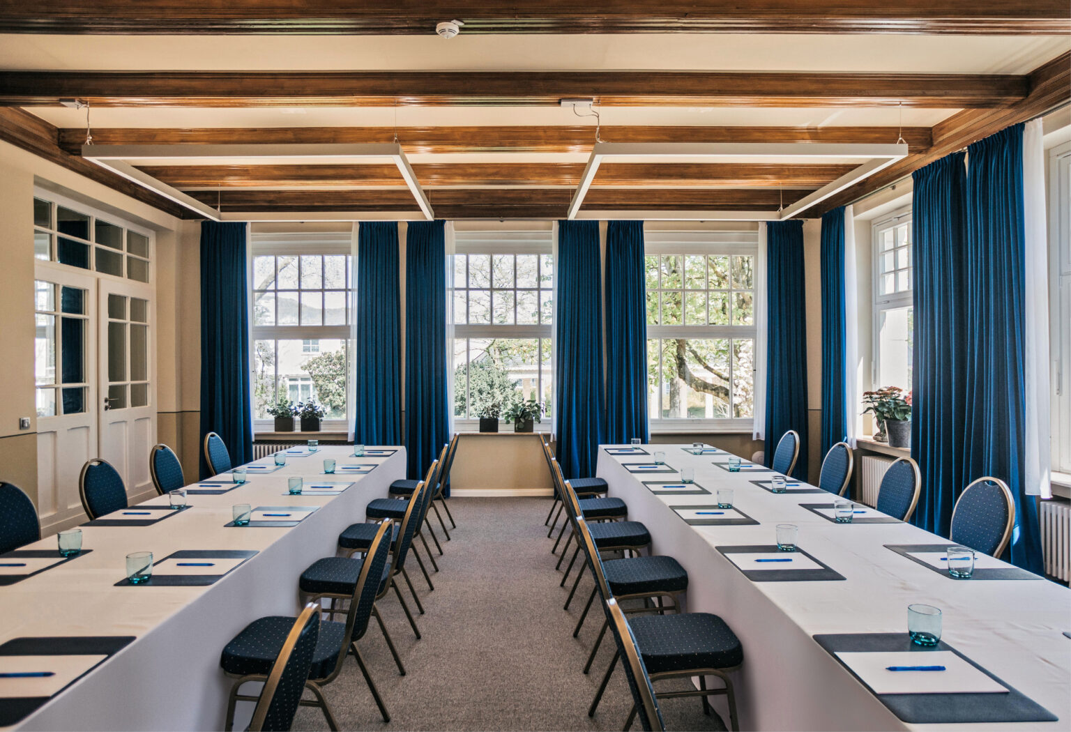 Konferenzraum mit U-förmigen Tischen, blauen Stühlen, Notizblöcken und großen Fenstern mit blauen Vorhängen – ideal für Meetings oder Events mit Catering in Bad Honnef.