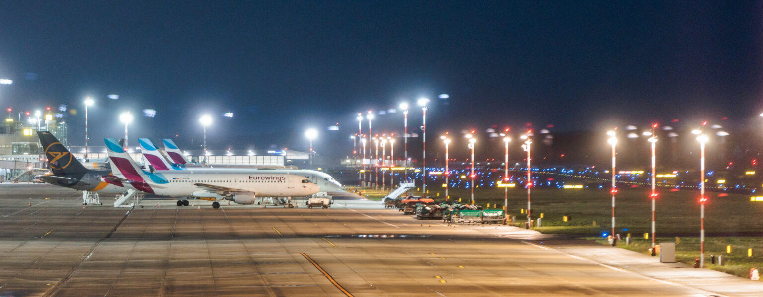 Nachts parken Flugzeuge an einem hell erleuchteten Terminal des Bahnhofs „Airport für Veranstaltungen“, in der Nähe befinden sich Bodenfahrzeuge.